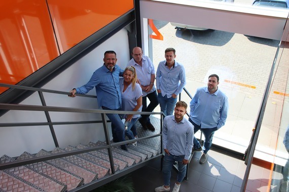 Sechs Personen stehen auf einer Treppe in einem modernen Innenraum mit orangefarbenen Akzenten.