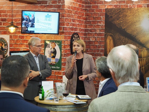 Eine Frau hält ein Mikrofon und spricht zu einer Gruppe von Menschen in einem Raum mit Backsteinwänden und einer großen Leinwand im Hintergrund.