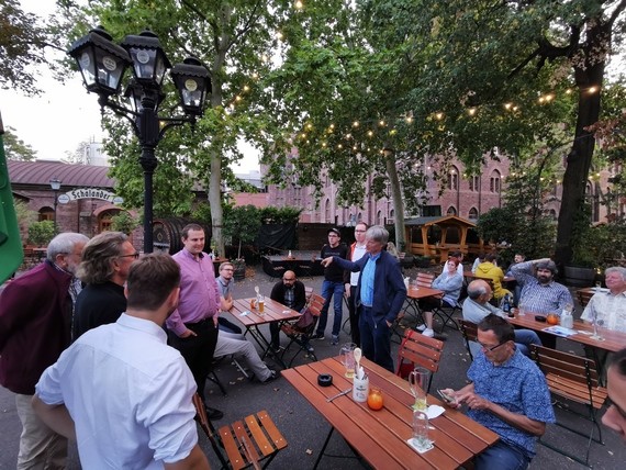 Gruppe von Menschen sitzt im Freien an Holztischen in einem belebten Außenbereich mit Biergartenatmosphäre.