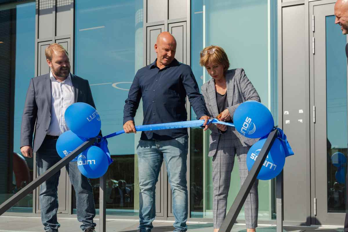 Drei Personen stehen vor einer Tür und schneiden gemeinsam einen blauen Scherentape, während blaue Luftballons im Hintergrund sichtbar sind.