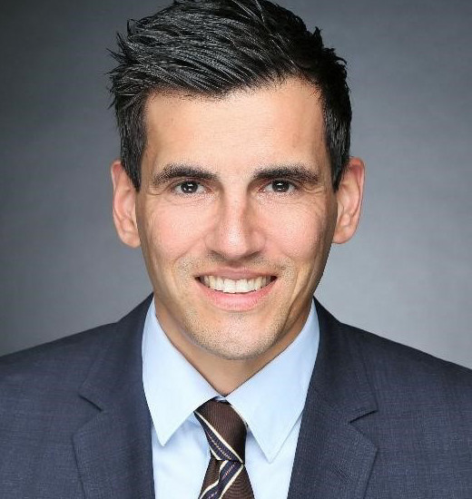 Ein Mann mit kurzen dunklen Haaren trägt ein dunkles Hemd mit Krawatte und steht vor einem unscharfen Hintergrund.