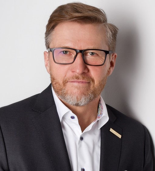 Ein Mann mit Brille, Bart und weißem Hemd unter einem dunklen Anzug steht vor einem unscharfen Hintergrund.