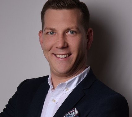 Ein Mann im Anzug mit Krawatte steht vor einem neutralen Hintergrund.