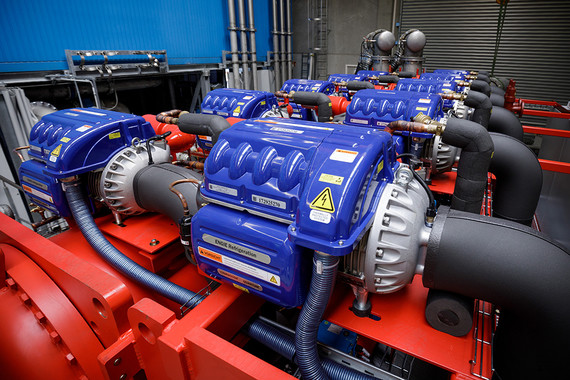 Drei blaue industrielle Kompressoren mit schwarzen Schläuchen und Warnetiketten stehen nebeneinander auf einem roten Gestell.