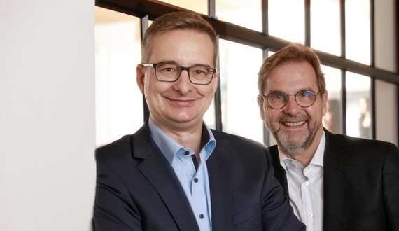 Zwei Männer in Anzügen stehen nebeneinander vor einem Fenster mit Gitter.