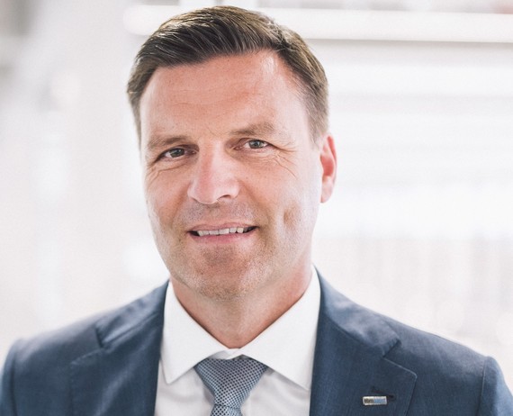 Ein Mann in einem Anzug mit Krawatte steht vor einem neutralen Hintergrund.