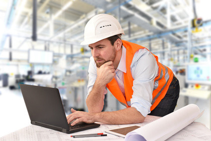 Ein Mann in Sicherheitsausrüstung arbeitet an einem Laptop in einer industriellen Umgebung.