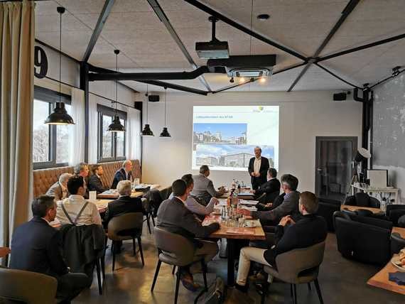 Eine Gruppe von Menschen sitzt in einem modernen Raum um einen Tisch, während eine Person an der Vorderseite eine Präsentation auf einem Bildschirm hält.
