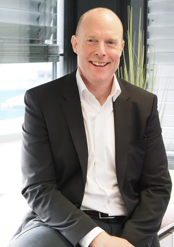 Ein Mann in einem Anzug sitzt lächelnd vor einem Fenster mit einem grünen Logo im Hintergrund.