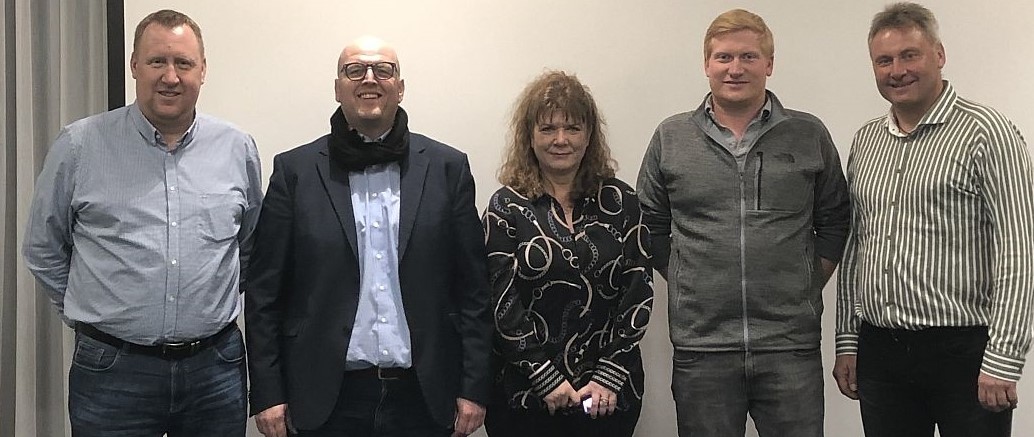 Fünf Personen stehen nebeneinander in einem Innenraum, alle lächeln und blicken in die Kamera.