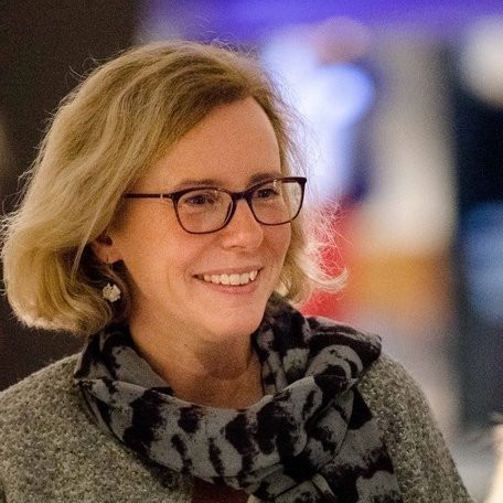 Eine Person mit kurzen blonden Haaren, Brille und einem gestreiften Schal lächelt in die Kamera.