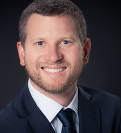 Ein Mann mit kurzen braunen Haaren und einem Lächeln trägt einen dunklen Anzug und eine Krawatte.