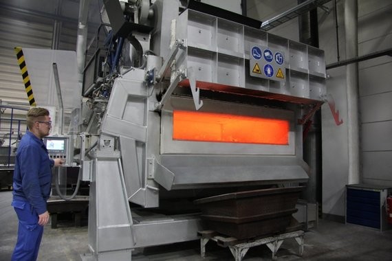 Ein Mann in Arbeitskleidung steht neben einem industriellen Ofen mit orangefarbener Glut im Inneren.