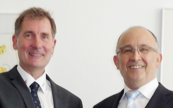 Zwei Männer in Anzügen und Krawatten stehen nebeneinander und lächeln.