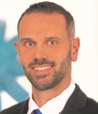 Ein Mann mit kurzen dunklen Haaren und einem blauen Hemd steht vor einem weißen Hintergrund.