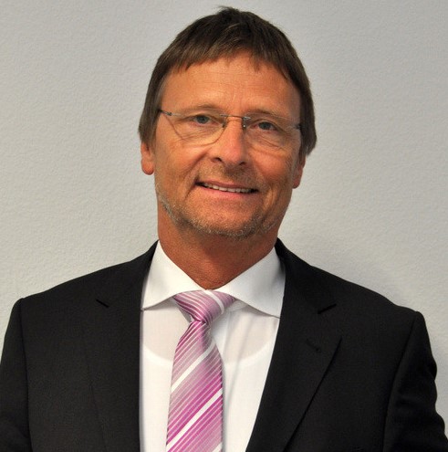 Ein Mann im Anzug mit Krawatte steht vor einem neutralen Hintergrund.