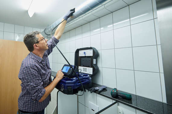 Ein Mann in einem karierten Hemd arbeitet in einer Fliesenwand, während er mit einem Messgerät an einer Rohrleitung hantiert.