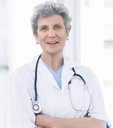 Eine ältere Frau in einem weißen Laborkittel mit Stethoskop steht lächelnd und hat die Arme verschränkt.