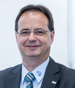 Ein Mann im Anzug mit Krawatte und Brille lächelt in die Kamera.