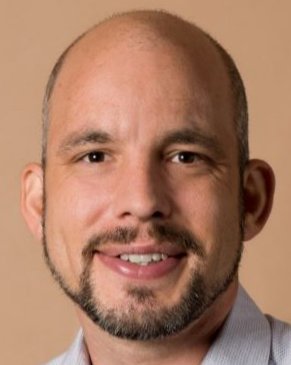 Ein Mann mit kahlköpfigem Haar, Bart und Lächeln auf einem neutralen Hintergrund.