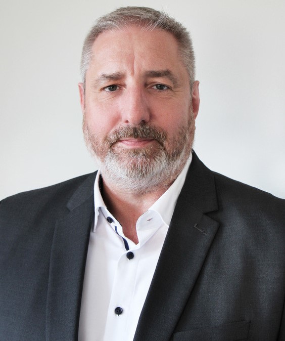 Ein Mann mit Vollbart und grauen Haaren trägt ein schwarzes Sakko über einem weißen Hemd.