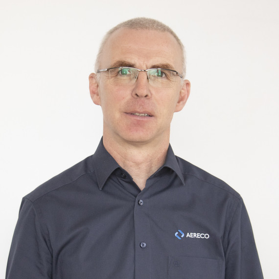 Ein Mann mit kurzen blonden Haaren und Brille trägt ein dunkelblaues Hemd mit einem Logo auf der Brust.