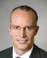 Ein Mann mit kurzen blonden Haaren und Brille trägt ein weißes Hemd und ein dunkles Sakko.