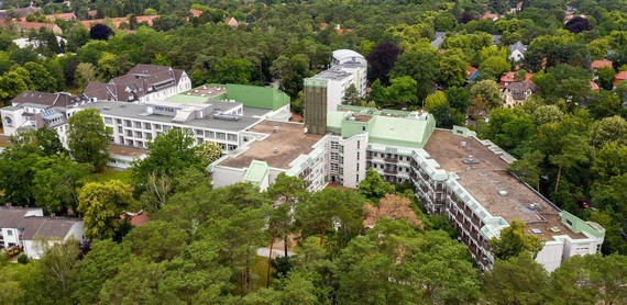 Ein Luftbild zeigt ein großes, mehrstöckiges Gebäude mit weißen Fassaden, umgeben von dichtem grünen Wald.