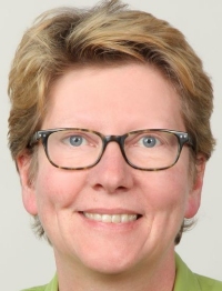 Eine Person mit kurzen Haaren, Brille und einem grünen Oberteil lächelt in die Kamera.