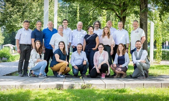 Gruppe von Menschen sitzt und steht im Freien vor Bäumen und einer Mauer.