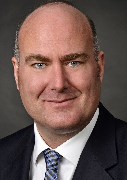 Ein Mann mit dunklem Haar und Bart trägt einen Anzug, ein weißes Hemd und eine karierte Krawatte.