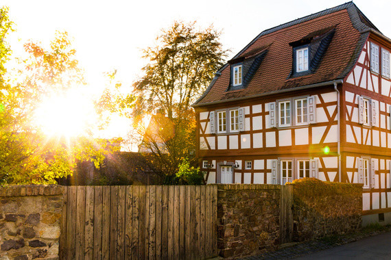 Ein Fachwerkhaus mit rotem Dach steht im warmen Sonnenlicht, umgeben von Bäumen und einer Mauer.