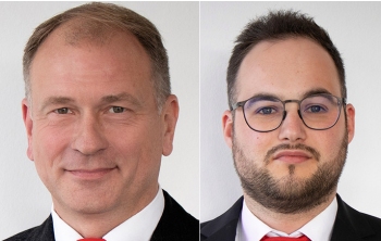 Zwei Männer in Anzügen und Krawatten, einer mit kurzen blonden Haaren und der andere mit kurzen braunen Haaren und Brille.