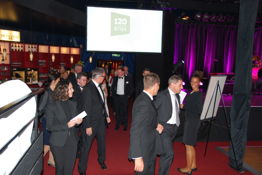 Eine Gruppe von Menschen in Anzügen steht auf einem roten Teppich vor einer Bühne mit einem Bildschirm, auf dem 