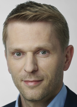 Ein Mann mit blondem Haar und blauem Hemd schaut direkt in die Kamera.