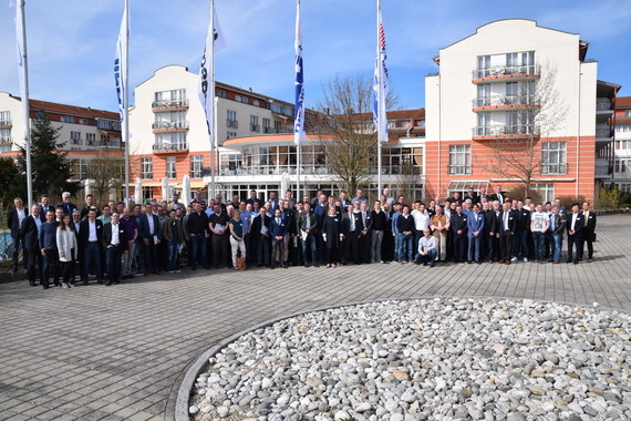 Gruppe von Menschen steht vor einem Gebäude mit Fahnen und einem gepflasterten Platz.