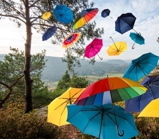 Ein buntes Regenschirmmeer schwebt über einem idyllischen Landschaftsblick mit Bäumen und einem klaren Himmel.