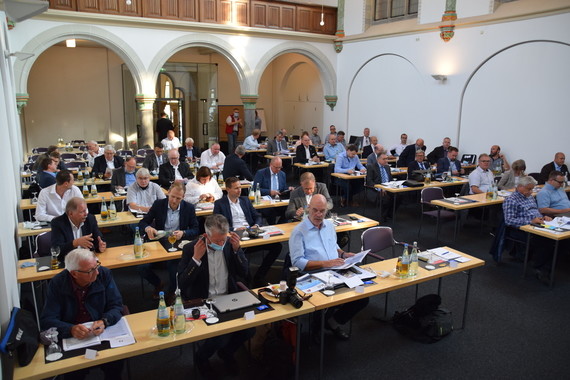 Eine Gruppe von Menschen sitzt in einem großen Saal an langen Tischen und nimmt an einer Veranstaltung oder Besprechung teil.