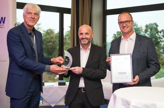 Drei Männer stehen nebeneinander, wobei einer einen Preis überreicht und einer ein Zertifikat hält.