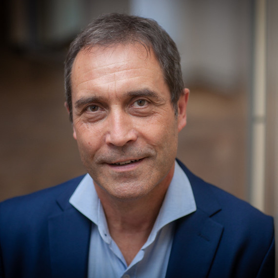 Ein Mann mit dunklem Haar und blauem Hemd sitzt in einem Büro oder Konferenzraum.