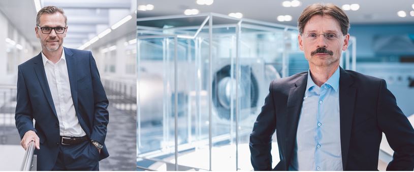 Zwei Männer in Anzügen stehen in einem modernen, industriellen Umfeld.