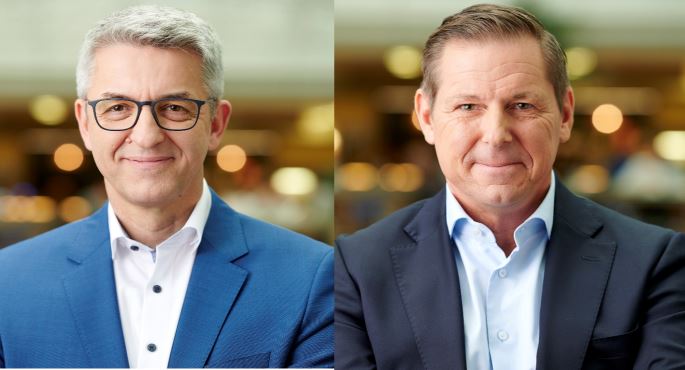 Zwei Männer in Anzügen und Hemden lächeln in die Kamera.