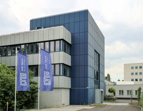 Ein modernes Bürogebäude mit blauen Fassadenplatten und hängenden Schildern, umgeben von Bäumen und einem klaren Himmel.