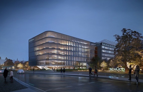 Ein modernes, beleuchtetes Bürogebäude steht am Abend an einer belebten Straße mit Bäumen und Passanten.