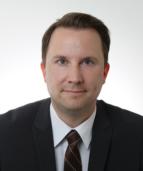 Ein Mann in Anzug und Krawatte steht vor einem unscharfen Hintergrund.