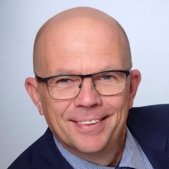 Ein Mann mit kahlen Haaren, Brille und lächelndem Gesicht trägt einen dunkelblauen Blazer über einem hellen Hemd.