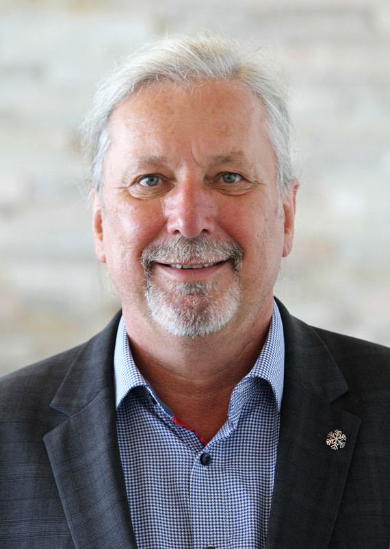 Ein Mann mit grauem Bart und Haaren trägt ein dunkles Sakko über einem hellblauen Hemd und steht vor einem neutralen Hintergrund.