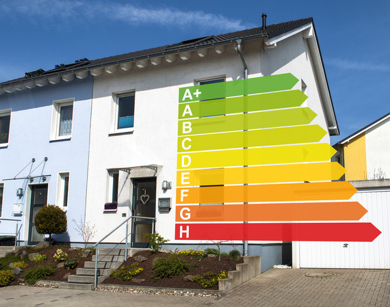 Ein Energieeffizienzdiagramm mit den Kategorien A+ bis H steht vor einem weißen Haus.