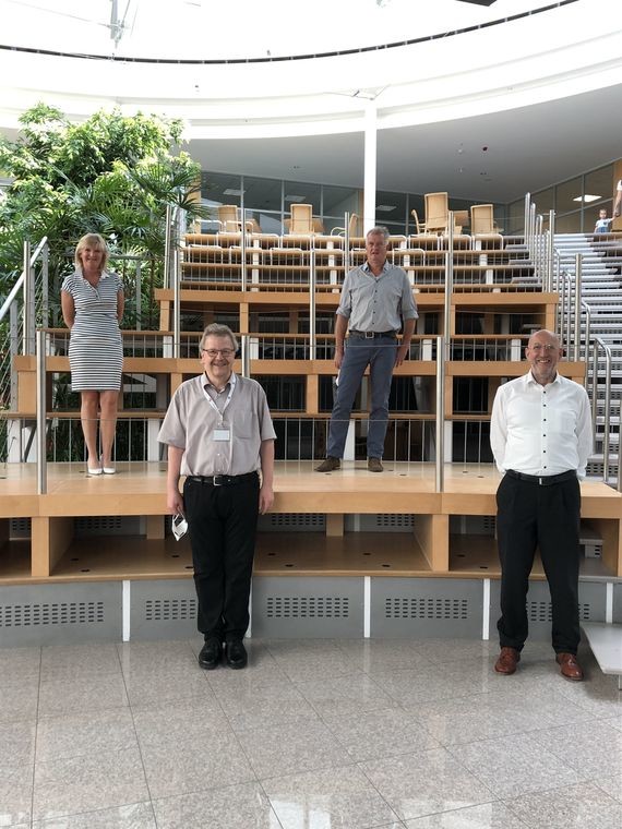 Vier Personen stehen in einem modernen, hellen Raum mit mehreren Ebenen und Bänken im Hintergrund.