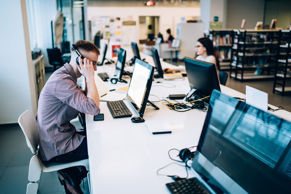 Mann sitzt an einem Tisch in einem Büro und telefoniert, während andere Personen im Hintergrund arbeiten.